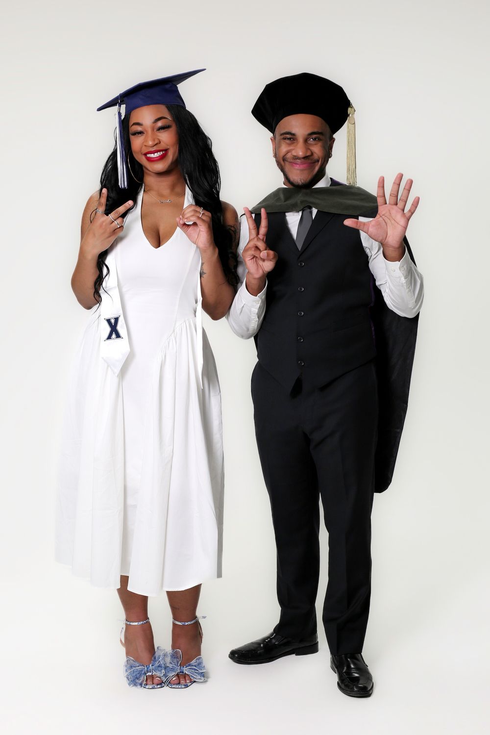 Two graduates in caps and gowns pose together, smiling and gesturing on a white background.