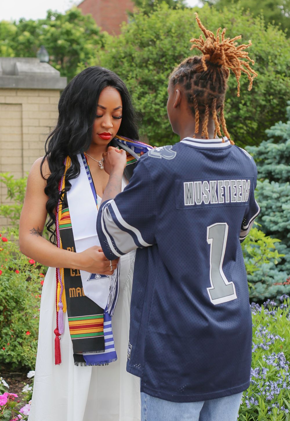 Graduate in white dress with colorful stole poses with person in Musketeers jersey during outdoor graduation celebration.
