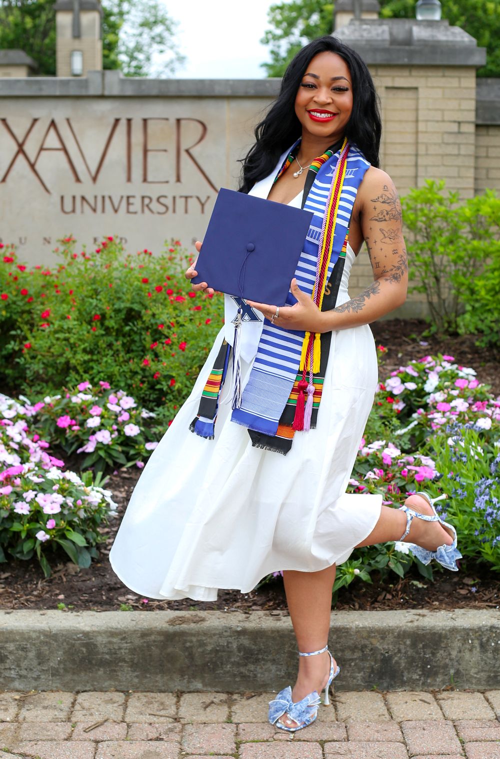 Woman in white dress holding graduation cap and diploma in front of Xavier University sign with colorful kente stole.
