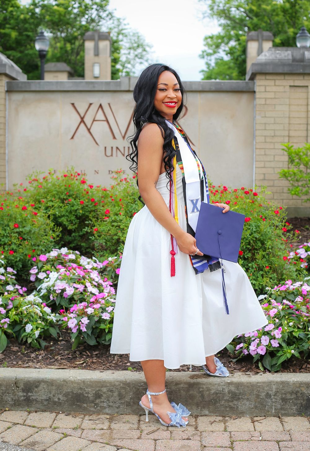 Graduate in white dress with kente stole poses proudly with navy cap in front of Xavier University entrance sign.