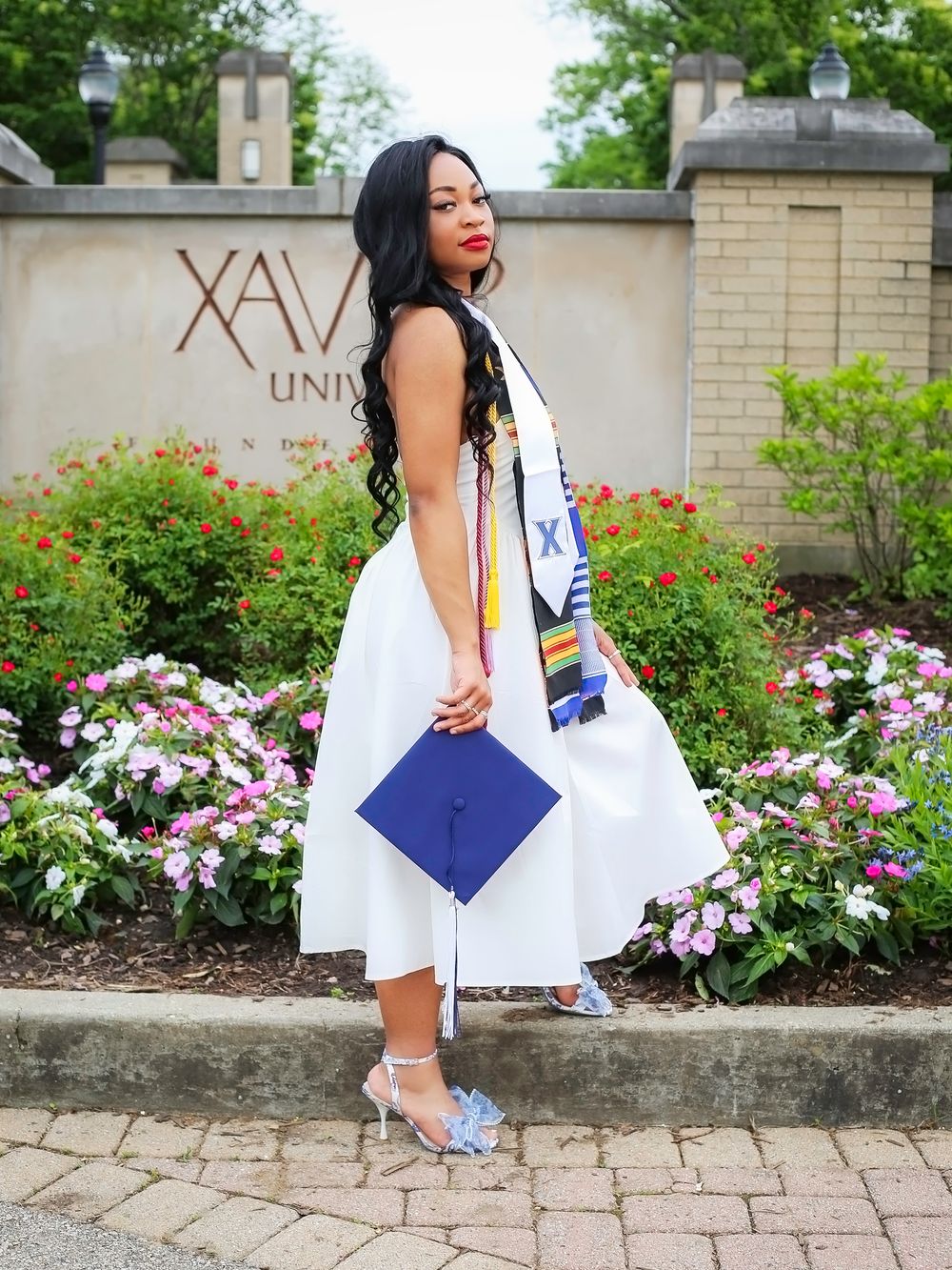 A woman in a white dress holds a blue graduation cap in front of Xavier University sign, wearing a colorful stole.