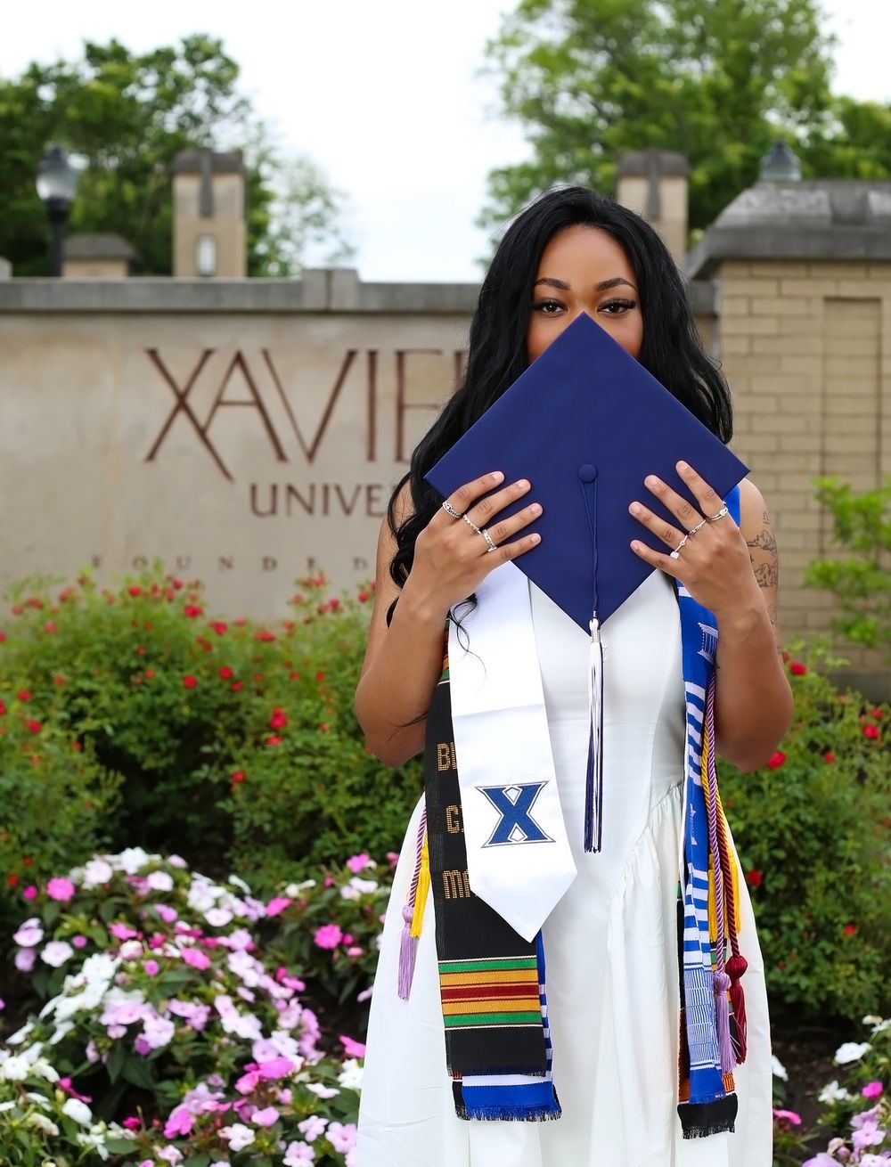Xavier University graduate in white gown holds navy cap, adorned with colorful stoles, posing in front of campus sign.
