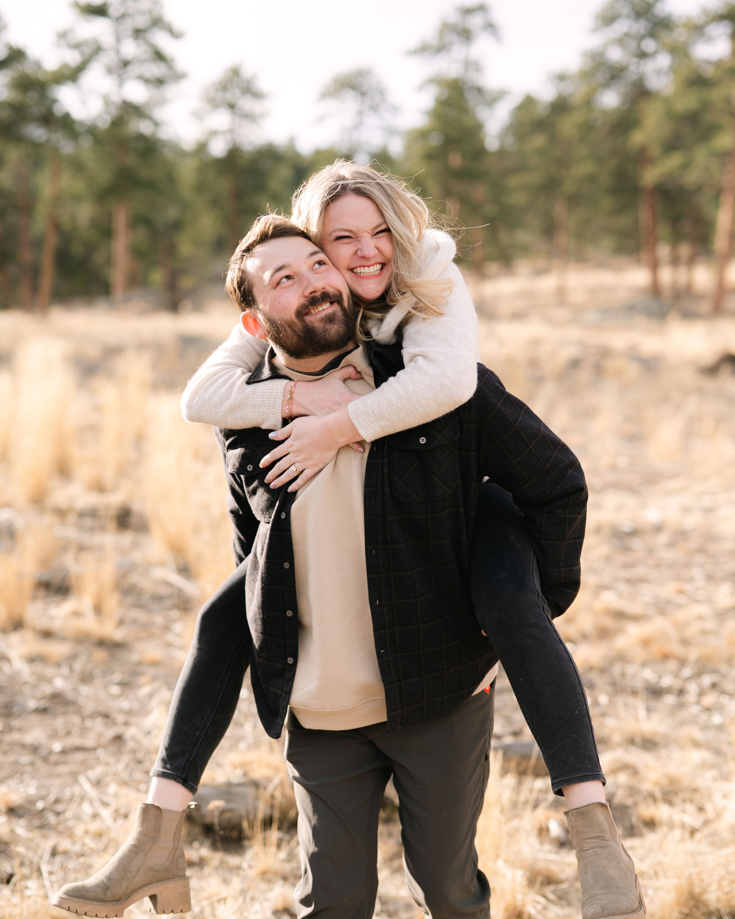 A playful couple shares joyful moments in a rustic outdoor setting with golden light and pine trees in the background.