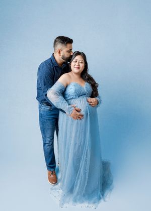 Couple in matching blue outfits posing for maternity photo, man kissing pregnant woman's cheek in studio.