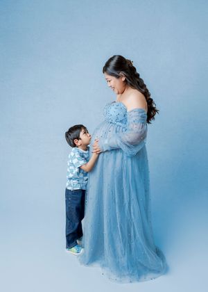 Pregnant woman in blue tulle gown smiling at toddler son touching her baby bump in studio.