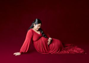 Pregnant woman in red gown sitting gracefully against red backdrop, cradling baby bump in elegant maternity photo.