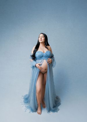 Pregnant woman in blue tulle maternity gown cradling baby bump against soft blue studio background.