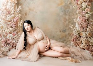Pregnant woman in beige tulle gown poses gracefully among pink floral arrangements in a dreamy maternity portrait.