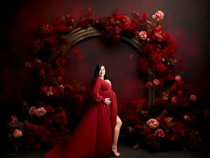 Pregnant woman in red gown poses before floral arch with red roses in elegant maternity photography studio portrait.