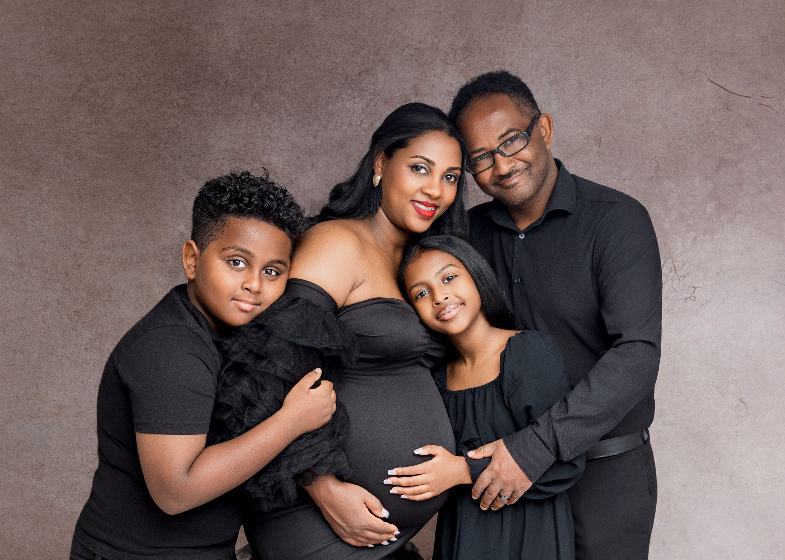 A family wearing coordinated black outfits poses together for a maternity portrait against a textured brown backdrop.