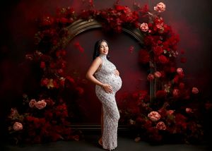 Pregnant woman in lace gown poses before floral arch with red roses for elegant maternity portrait.