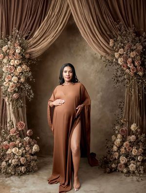 Pregnant woman in elegant brown maternity gown poses before floral arch with roses and draped curtains.