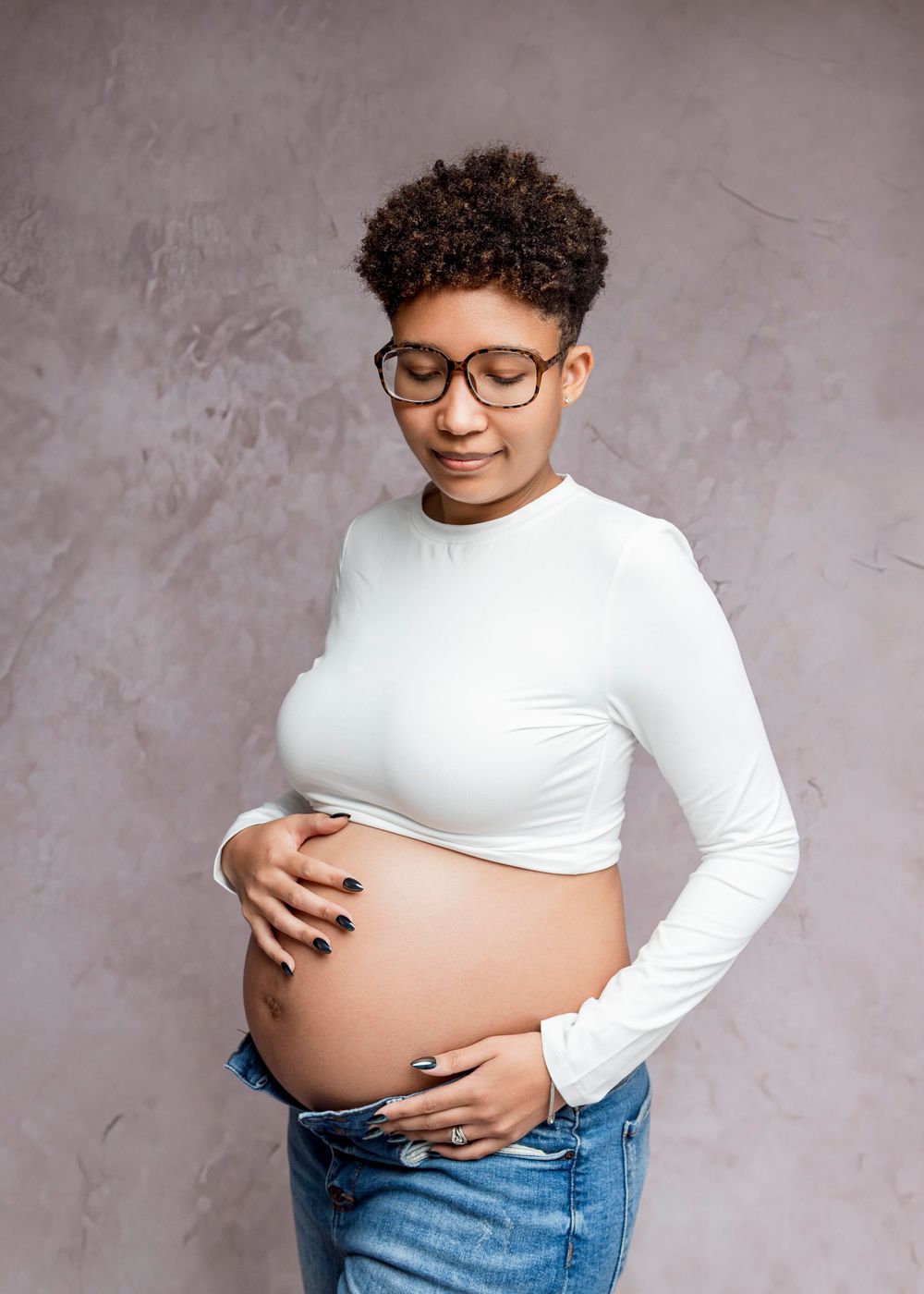 A pregnant person in a white long sleeve top and jeans poses against a gray backdrop in a progression of photos.