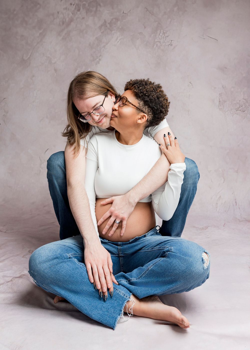 A couple shares an intimate moment while sitting on the floor against a textured gray backdrop.