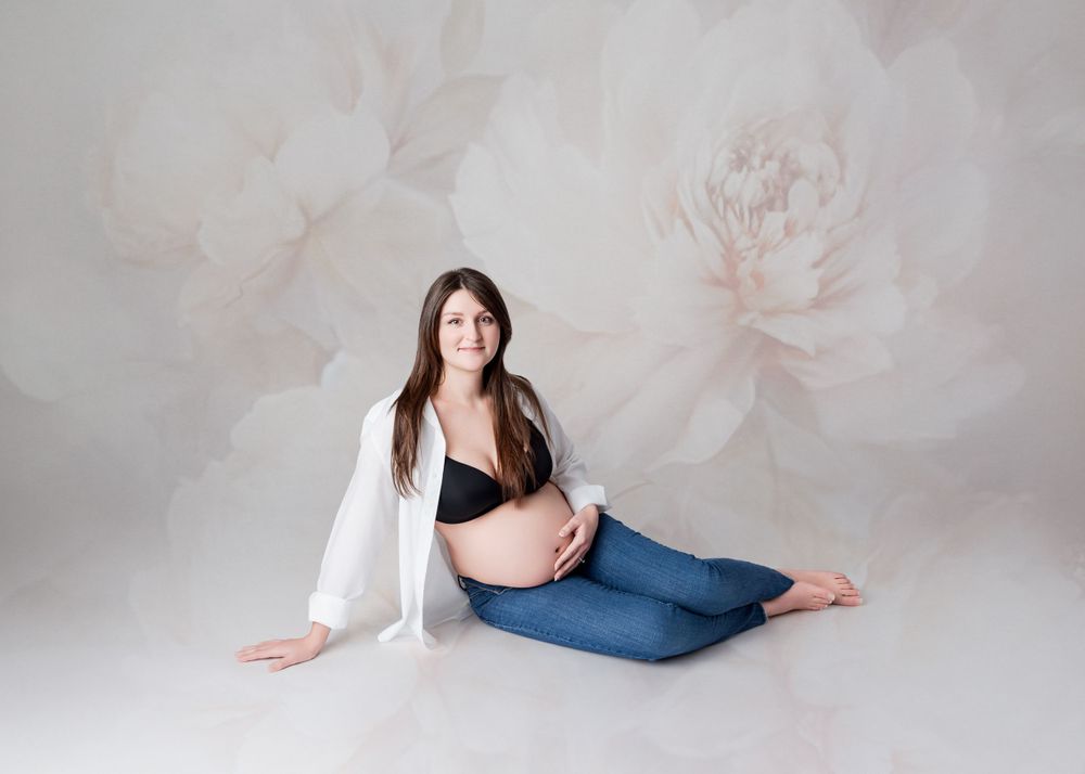 Expectant mother in white cardigan and jeans poses on floor against floral background during maternity photoshoot.