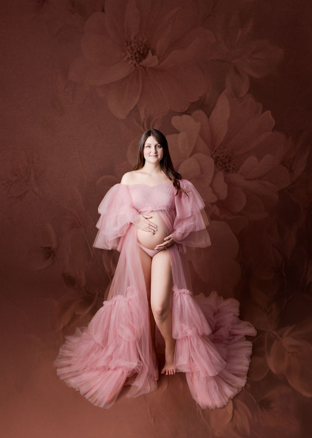 Ethereal maternity portrait featuring a flowing pink tulle robe against a floral burgundy backdrop.