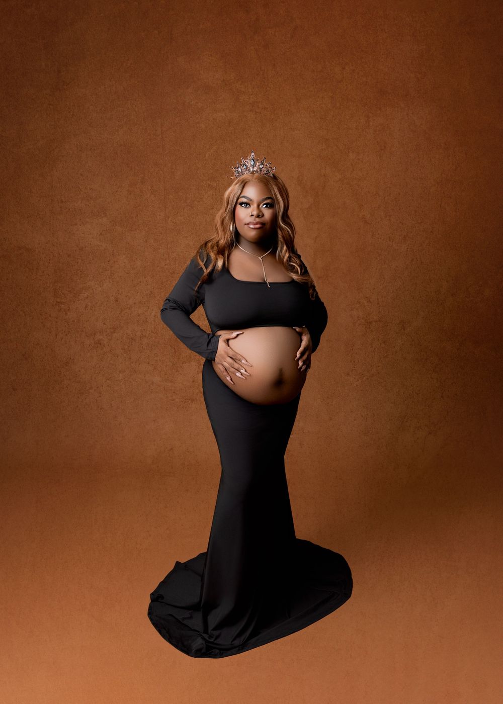 Elegant maternity portrait of an expectant mother in a black formal gown and tiara against a brown backdrop.