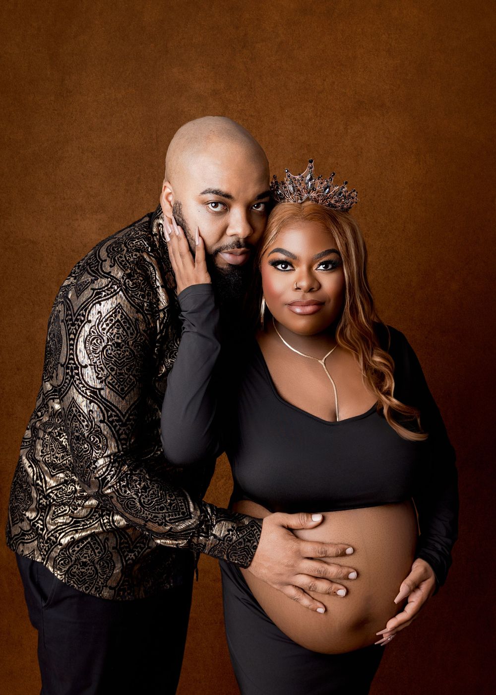 Elegant maternity photo against brown backdrop featuring couple in black and gold formal wear with woman wearing tiara.
