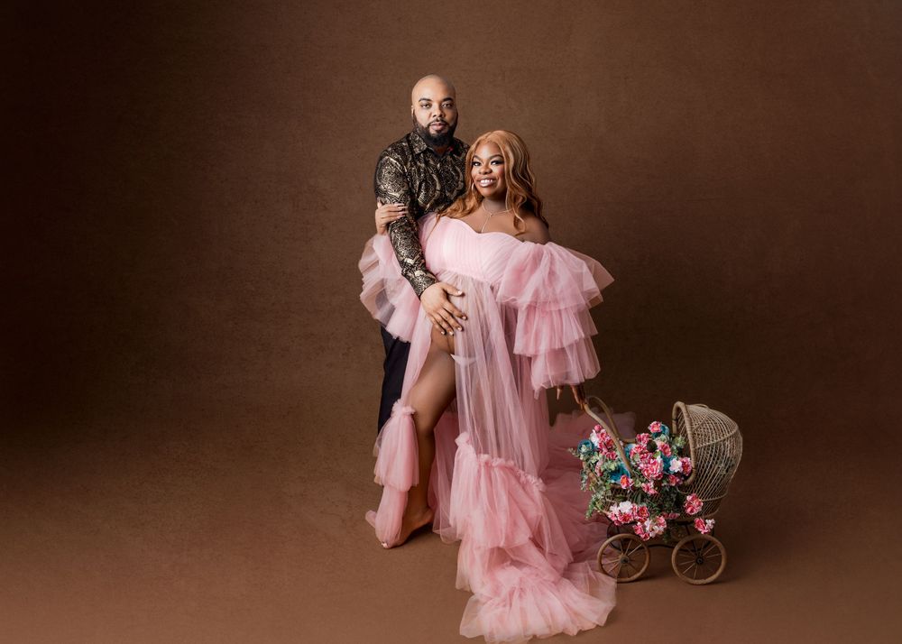 Studio portrait featuring elegant pink tulle gown and vintage pram with floral arrangement against brown backdrop.
