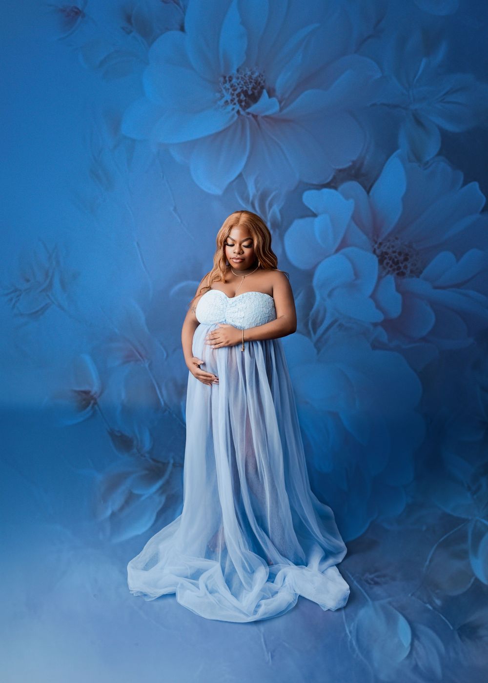 Ethereal maternity photo with flowing light blue gown against floral backdrop in blue tones.