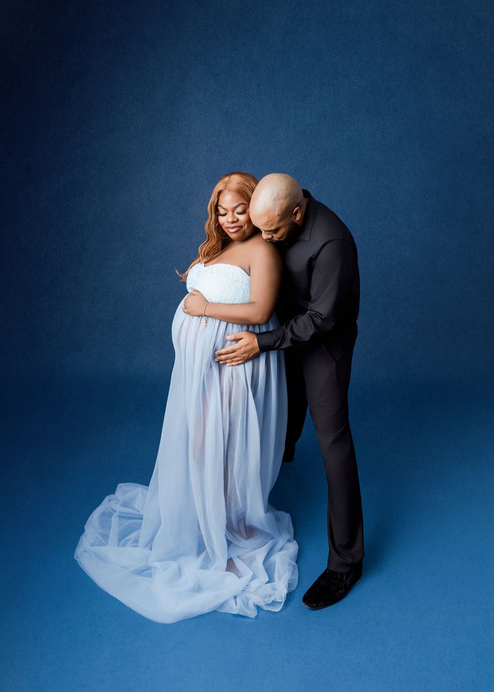 Intimate maternity photo of couple embracing against dark blue backdrop, woman wearing flowing white gown.