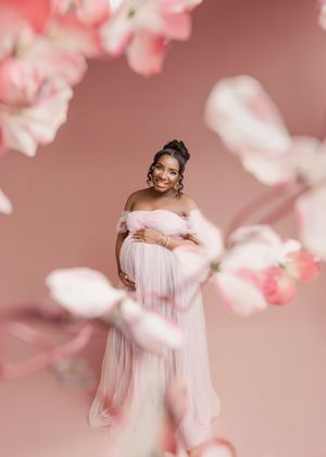 A dreamy maternity photoshoot with soft pink cherry blossoms and flowing white tulle dress creating an ethereal atmosphere.