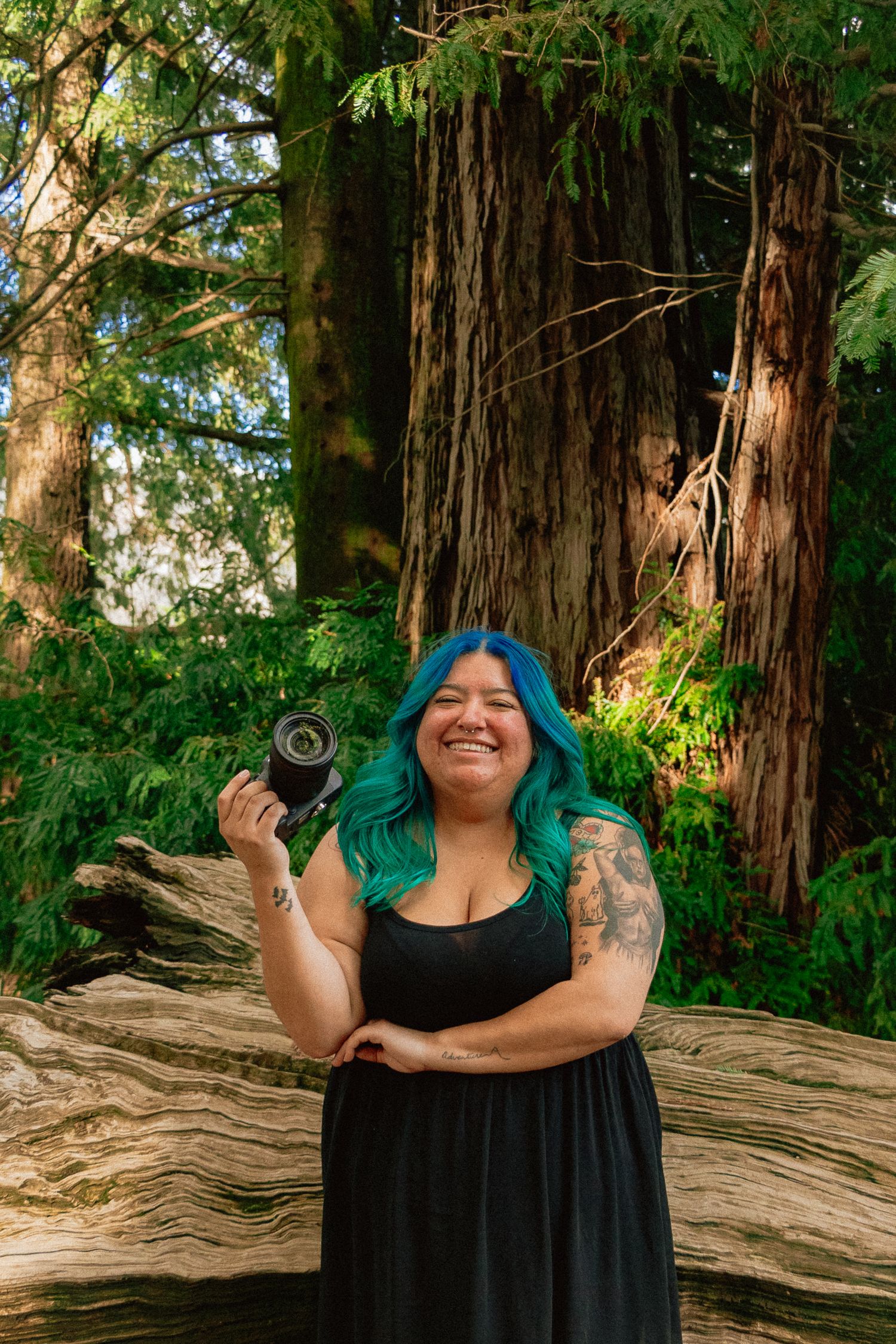 A woman photographer with vibrant blue and green hair wearing black dress smiles joyfully in front of towering redwood trees.