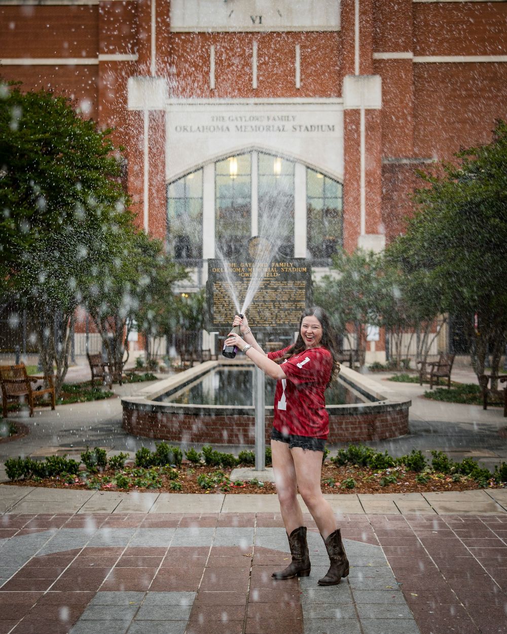 Woman in red shirt and cowboy boots sprays champagne in front of Oklahoma Memorial Stadium fountain.