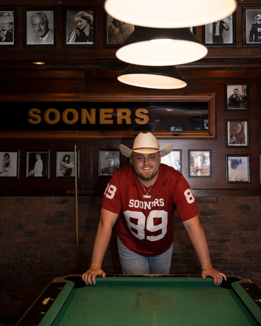 A pool player in a Sooners jersey leans over a green billiards table under pendant lights with vintage photos on wood walls.