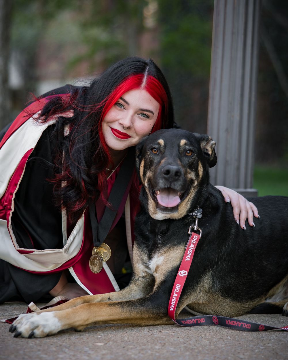 A happy black and tan dog wearing a red leash poses with a companion in casual gothic attire outdoors.