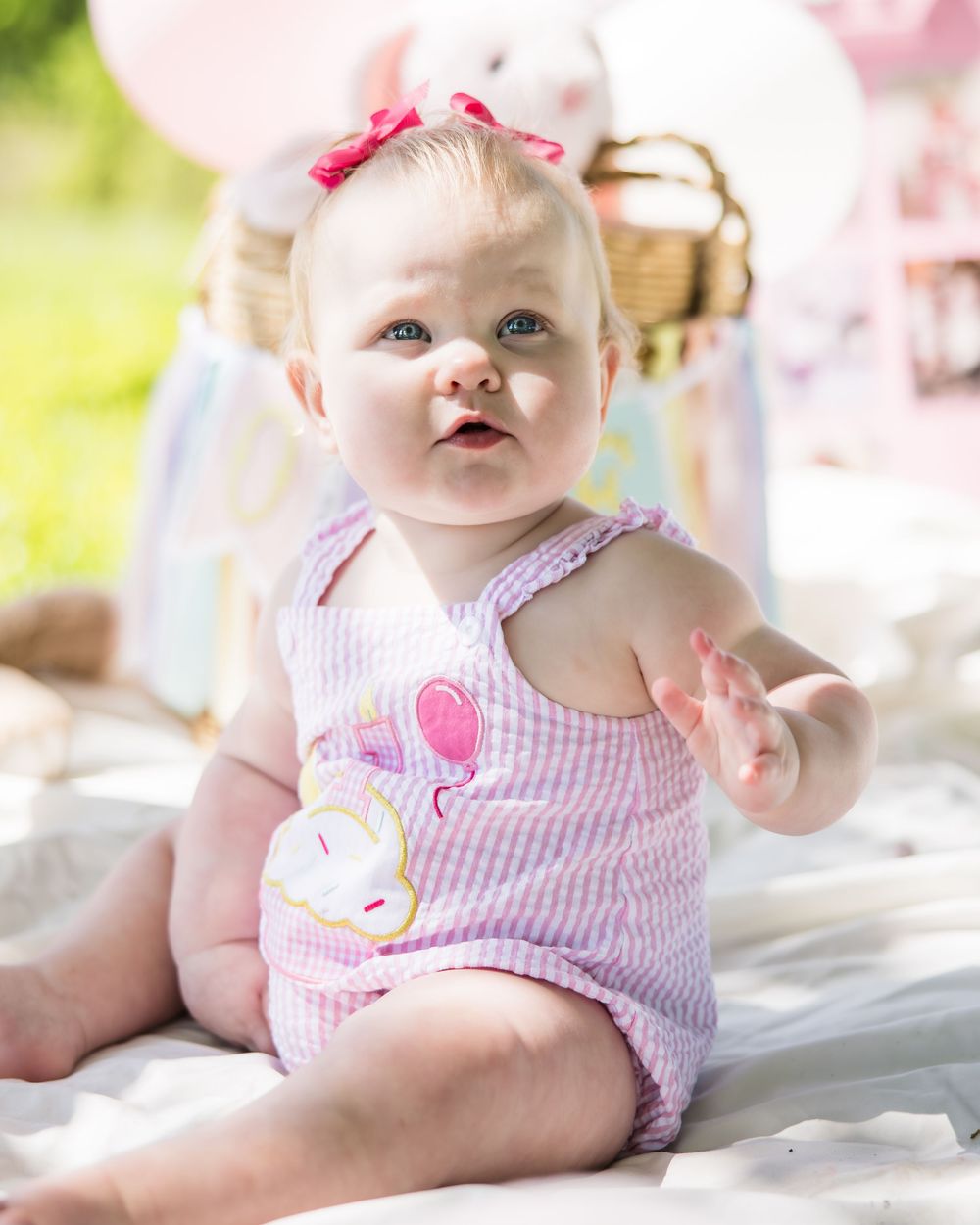 Baby girl in pink striped romper with bunny applique sits on blanket outdoors at a birthday party celebration.
