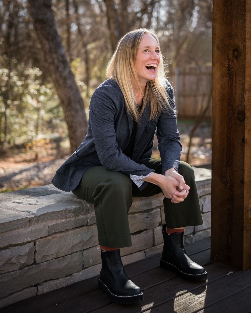 Smiling blonde woman sitting on stone wall outdoors, wearing navy blazer, green pants, and black boots.