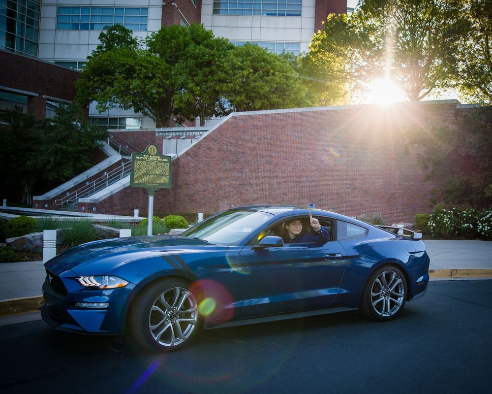 Blue Ford Mustang sports car parked on a curved driveway with sunlight flare and modern building in background.