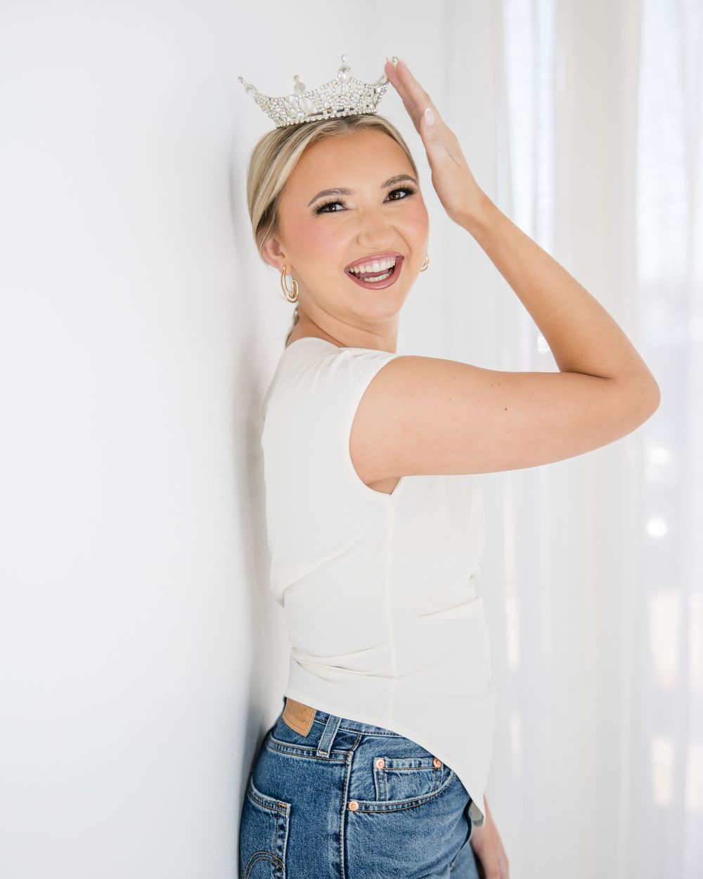 A person in a white tank top and blue jeans poses playfully against a white wall in a bright studio setting.