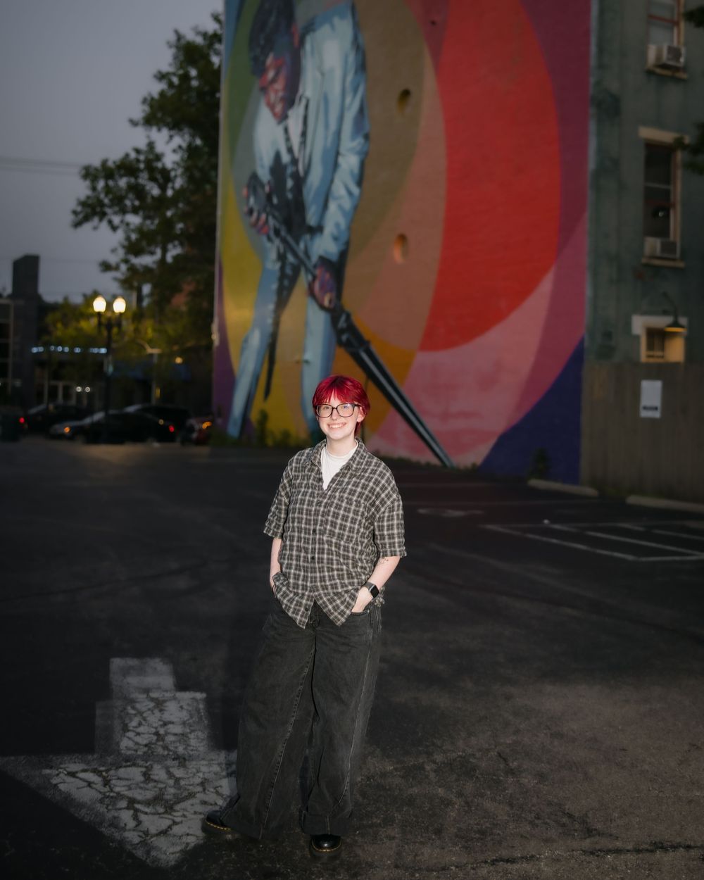 Person with red hair stands in parking lot before colorful urban mural at dusk.