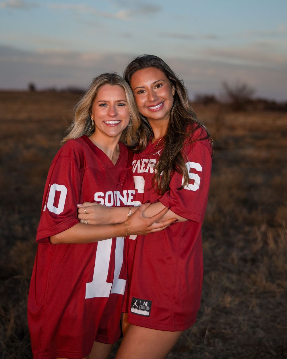 Two friends in matching red sports jerseys share a joyful moment together during sunset in an outdoor field setting.