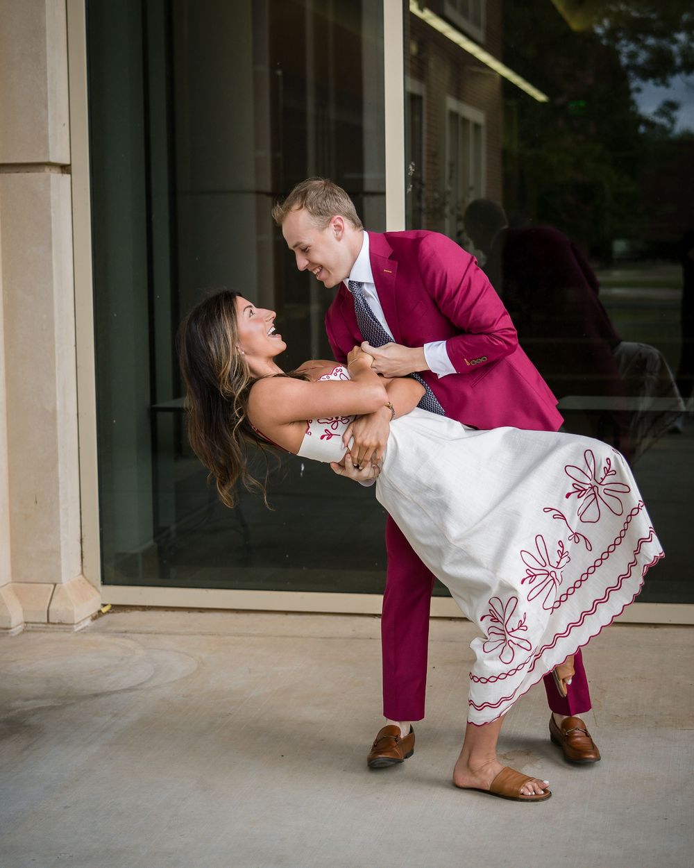 Couple dancing outdoors, man in magenta suit dipping woman in white embroidered dress, smiling joyfully.