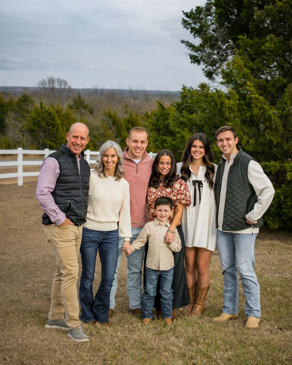 A group of people standing together outdoors in casual clothing against a rural backdrop with white fencing.