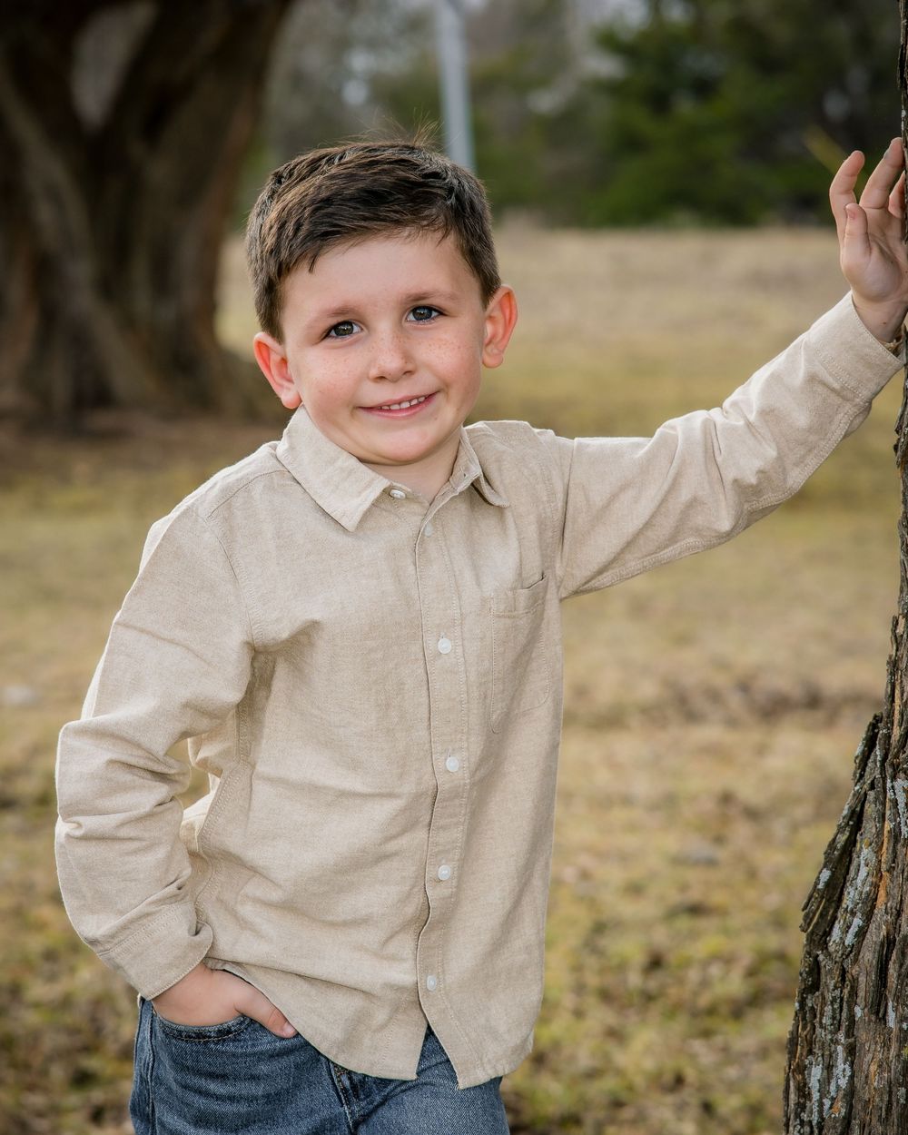 A young child in a beige button-down shirt and jeans poses playfully next to a tree stump outdoors.