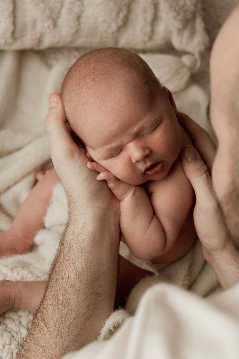 Sleeping newborn baby cradled gently in hands against a cream textured blanket background.