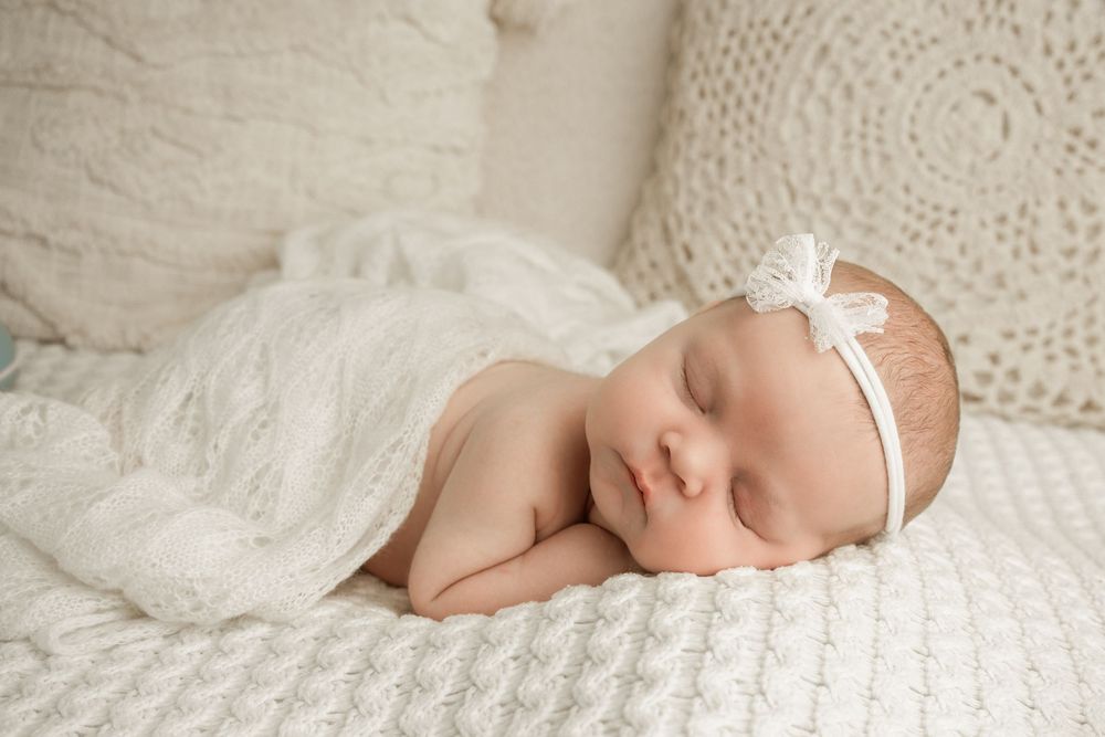 Sleeping newborn baby wrapped in soft white lace blanket lying on textured fabric in natural light.