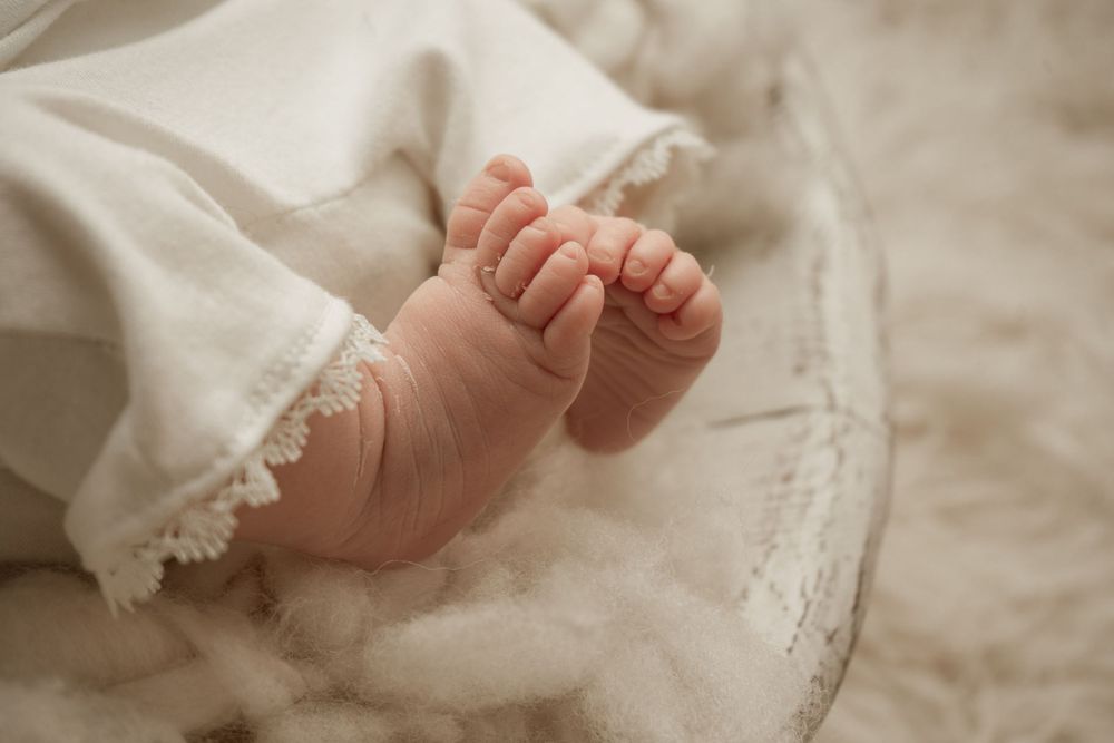 Close-up photo of tiny newborn baby feet wearing a delicate white lace dress against a soft cream background.