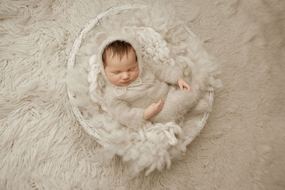 Peaceful sleeping newborn wrapped in soft white fabric lying in round basket on textured beige carpet.