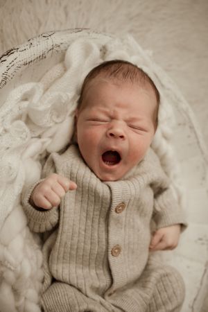 A yawning newborn baby wearing a beige knit sweater lying on soft white blankets.