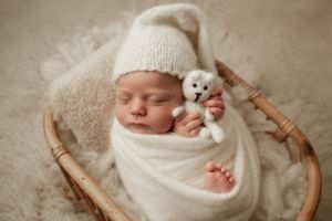 Sleeping newborn wrapped in white blanket with small stuffed animal in rustic basket.