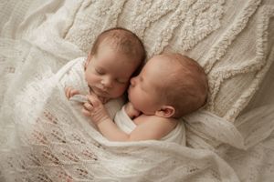 Two peacefully sleeping newborn babies snuggle together on soft white lace fabric in a gentle, tender moment.