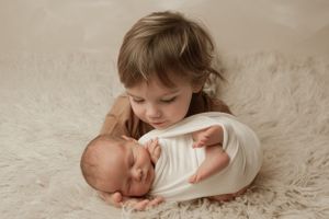 Two young children in white clothing cuddle together on a soft fluffy cream-colored rug.
