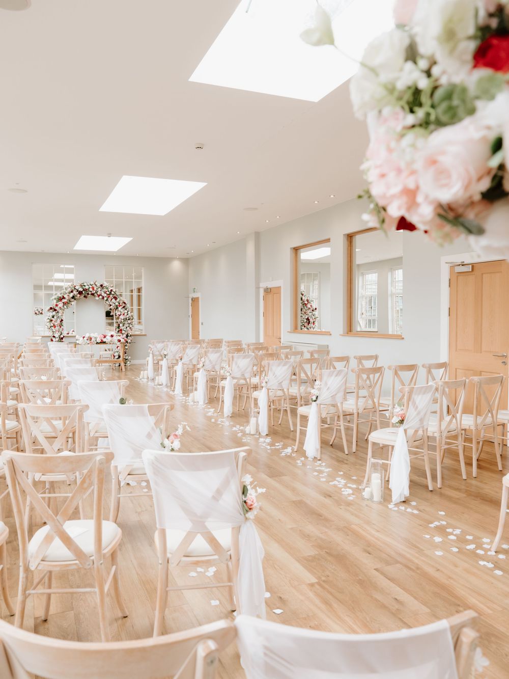 Light and airy indoor wedding ceremony venue with white crossback chairs and floral arch decor on hardwood floors.