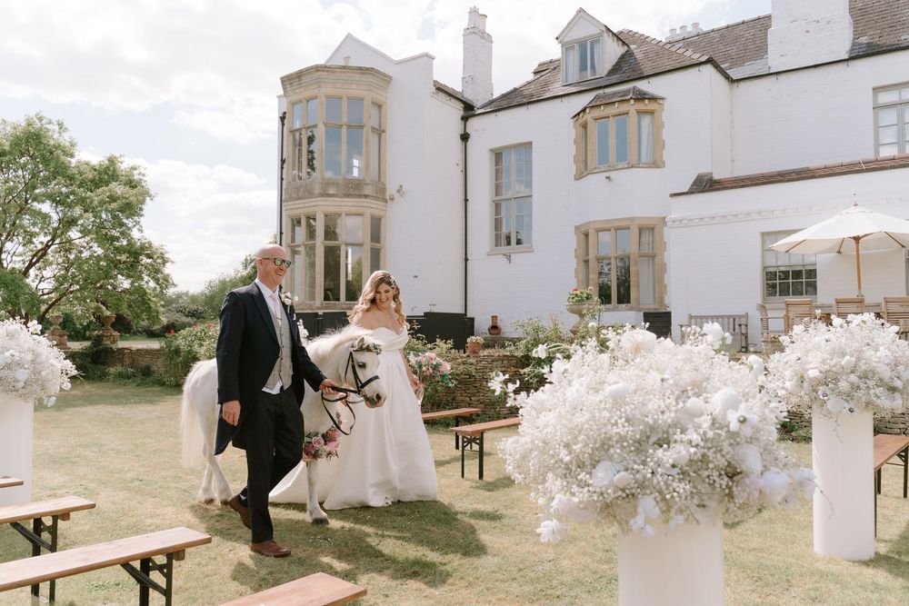 A couple stands next to wooden benches adorned with white floral arrangements outside an elegant white stone manor house.