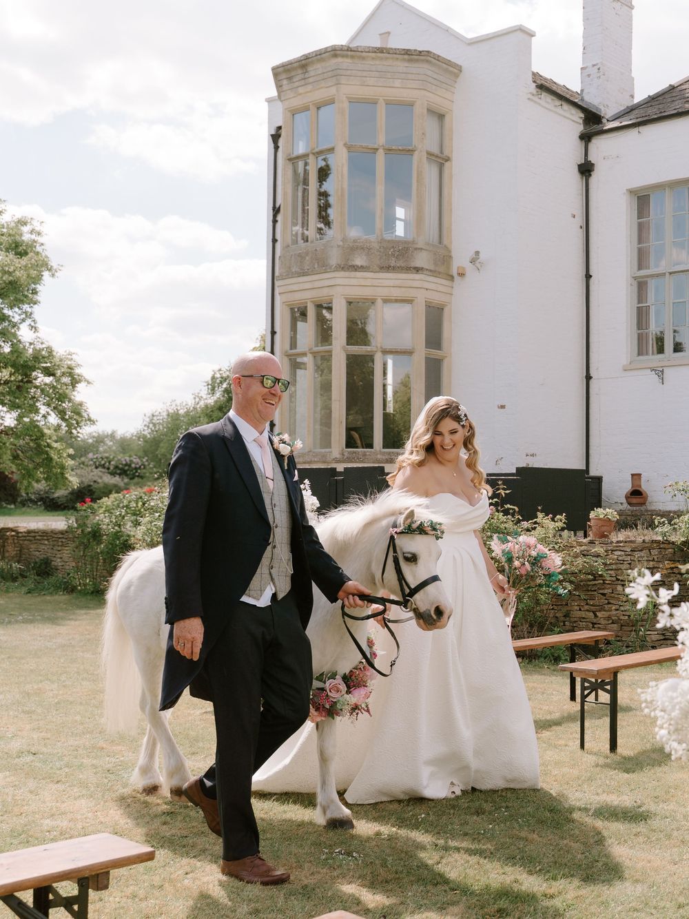 A couple at their wedding celebration poses with a white horse outside a stately stone building with a bay window.